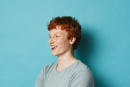 Young man with red hair and freckles smiling and looking to the side wearing gray t shirt on blue background. Casual portrait of male with happy expression and relaxed posture.の写真素材