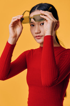 Young woman in red sweater holding sunglasses in front of yellow background. Asian female model with long dark hair posing for fashion portrait with casual expression and minimal makeup.の写真素材