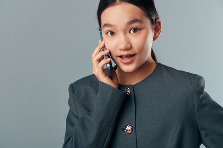 Young woman talking on mobile phone with surprised expression wearing gray blazer isolated on gray...