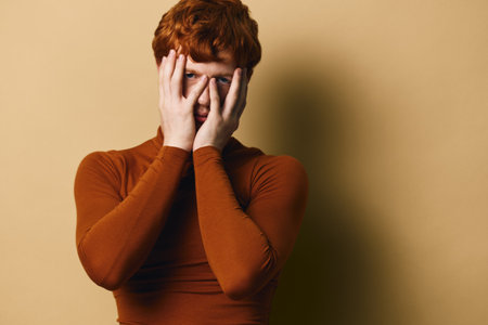 Redhead man in brown turtleneck covering face with hands looking through fingers, portrait on beige...