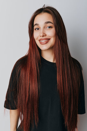 Young woman with long brown hair smiling and posing in black t shirt on light gray background....