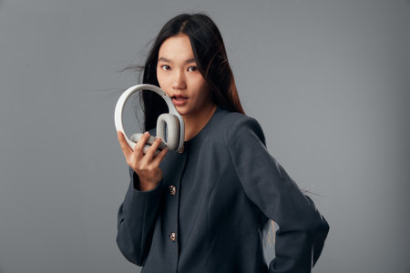 Young woman holding headphones with one hand looking at camera on grey background. Portrait of female model with long hair wearing dark jacket for music or technology concept.の写真素材
