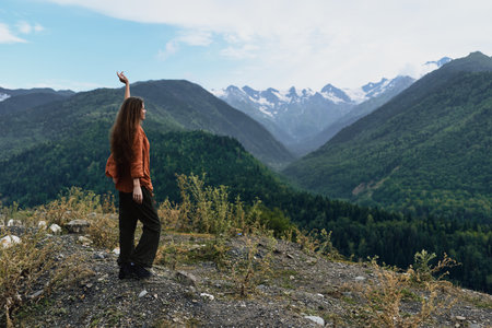A hiker stands on rocky ground amid lush hills and towering snowy peaks, raising one arm toward the expansive valley for a dramatic outdoor landscape momentの写真素材