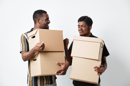 Two black men with dark skin carrying cardboard boxes and smiling while moving or working together in casual clothes on white background for teamwork and delivery.の写真素材