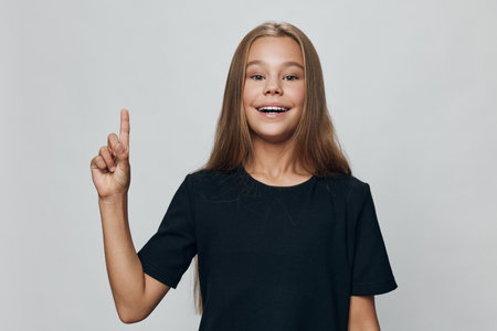 Girl with long hair wearing black t-shirt smiling and pointing up with forefinger against neutral...