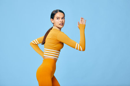 Young woman in orange fitness outfit posing in a running stance against a solid light blue studio...
