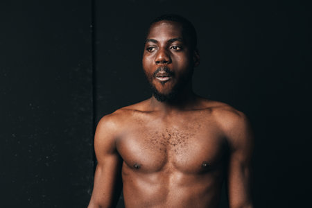 African man shirtless with muscular body and beard in creative conceptual visual style, dark background, portrait with serious expression and natural lighting.の写真素材