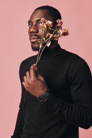 African man in black turtleneck holding dried flowers near face with creative conceptual visual style on pink background. Portrait with thoughtful expression and artistic mood.の写真素材