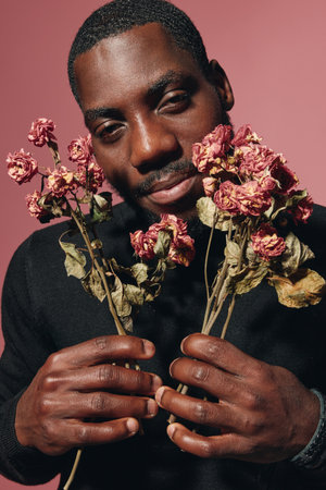 African man holding dried roses with closed eyes in creative conceptual visual style, portrait with dark skin and emotional expression on pink background for artistic photography.の写真素材