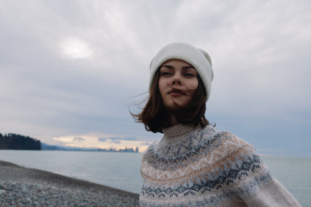Woman wearing a knit sweater and beanie by the seaside, wind in hair, calm water and cloudy sky, casual winter fashion portrait with outdoor coastal moodの写真素材