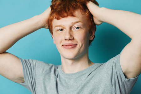 Young man with red hair wearing a gray t shirt smiling and holding hands behind head against blue background. Portrait of confident male with casual pose and natural expression.の写真素材