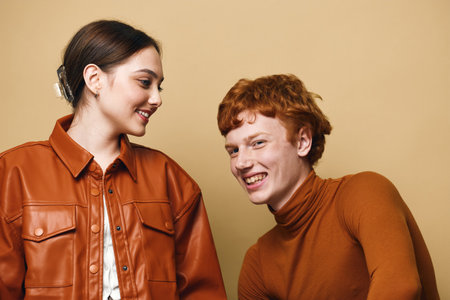 Two young people with red and brown hair wearing brown clothing smiling and interacting on beige background in casual indoor portrait studio lighting.の写真素材