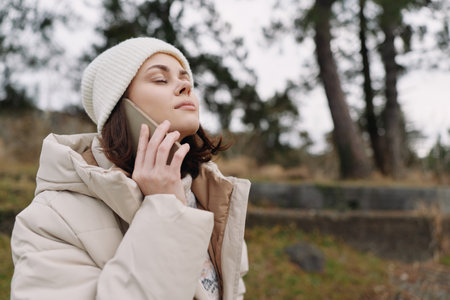Woman outdoors wearing a coat and beanie talking on a phone in a tranquil park, capturing a candid...