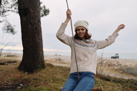 Woman on a swing outdoors, wearing a cozy sweater and beanie, smiling as she enjoys nature near the beach. Casual leisure scene with trees and open sky, capturing a playful mood.の写真素材