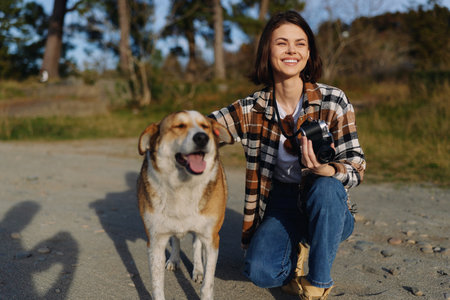 Woman kneels beside a friendly dog on a sunny outdoor park path, capturing memories with a camera and enjoying companionship with her pet on a bright day.の写真素材