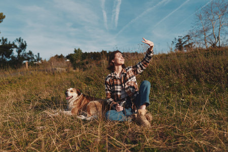 Woman and dog in an open field under a blue sky, enjoying nature and sunshine. Casual plaid shirt, jeans and boots as she sits and waves toward the bright horizon.の写真素材