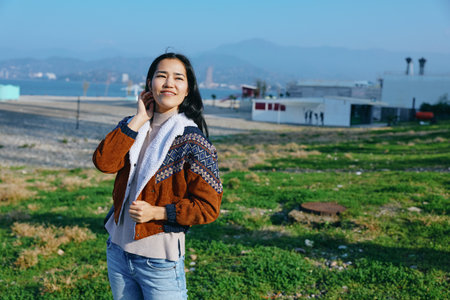 Smiling woman in a knitted jacket and scarf stands on a grassy coastal field with sea and hills in...