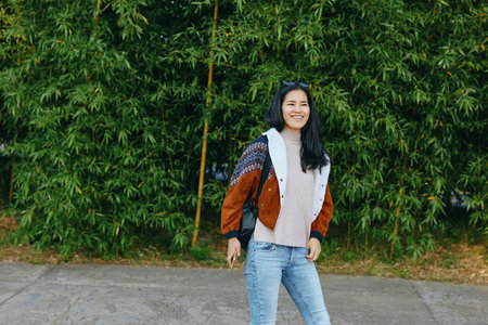 Smiling woman stands on a sidewalk beside a lush green hedge, wearing a color block jacket and denim jeans, capturing a casual street fashion moment with relaxed style and sunny outdoor vibe.の写真素材