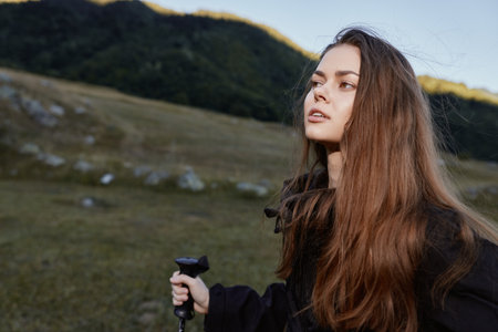 A young woman stands outdoors in a rugged landscape, gripping a trekking pole and gazing into the distance with calm confidence, capturing a mood of adventure.の写真素材