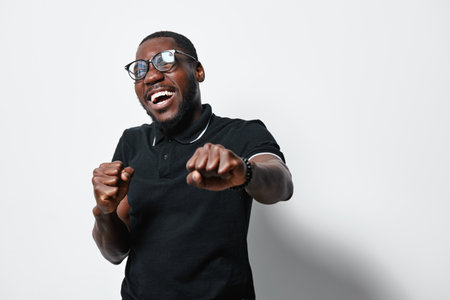 African American man with dark skin and glasses smiling and posing with fists up in black polo shirt on white background. Happy, confident and energetic male portrait.の写真素材