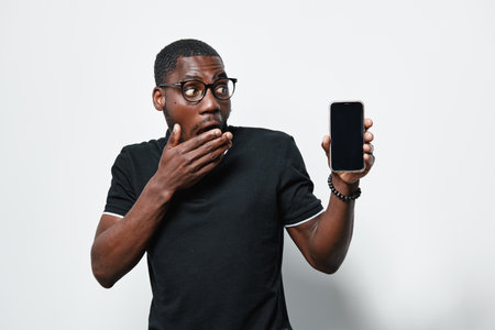 Black man with dark skin and glasses showing smartphone with blank screen in hand, surprised expression and casual black t shirt on white background for technology concept.の写真素材
