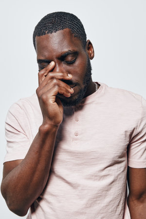 African American man with dark skin and short hair wearing light pink shirt standing with eyes closed and hand touching face in thoughtful or emotional pose on white background.の写真素材
