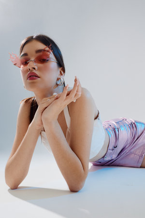 Young woman in trendy pink sunglasses and shiny shorts poses lying down on white floor. Studio shot...