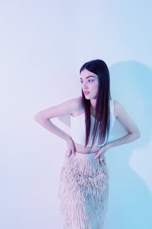 Young woman in a white crop top and textured skirt poses with hands on hips against a pastel...