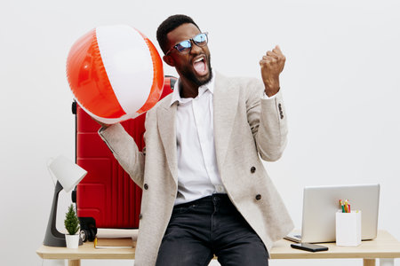 Excited cheerful young African-American man in smart casual clothing celebrating with a large beach ball in modern office environment, holding fist up in triumph, smiling, positive mood, lifestyle concept, studio shot.の写真素材