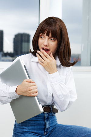 A surprised woman in casual attire holds a laptop while standing by a window, expressing curiosity and shock. The scene captures a moment of unexpected tech news or sudden realization.の写真素材