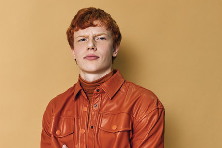 Young man with red hair wearing a brown leather jacket and turtleneck posing with crossed arms on plain background. Confident male portrait with casual fashion style and serious expression.の写真素材