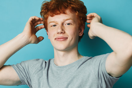 Young man with red hair and fair skin wearing grey t shirt posing with hands near head smiling on blue background. Casual style, confident mood and healthy appearance.の写真素材