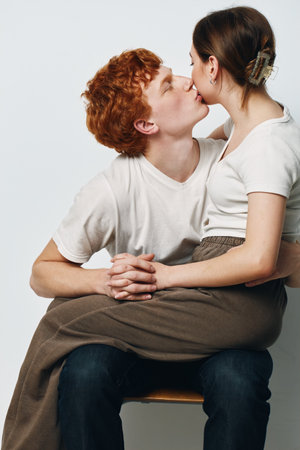 Couple in white shirts sitting on chair, young man holding girl on lap and kissing her cheek in intimate romantic moment with plain background.の写真素材