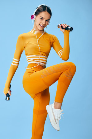 A woman in bright orange athletic wear energizes a dynamic workout with dumbbells, wearing headphones, lifting one knee for balance against a blue backdrop, showcasing fitness, sport, and activeの写真素材