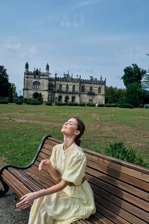 Woman on bench in park near historic building wearing yellow dress enjoying sunlight relaxation and...