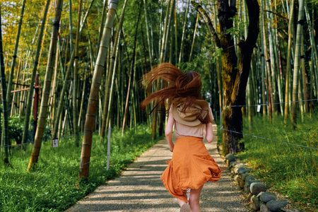 Woman running on a path through bamboo forest with backpack and orange skirt, rear view motion, sunlight filtering through tall stalks, outdoor trail adventure and carefree travel.の写真素材