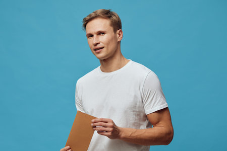 Confident young man with light brown hair wearing casual white t-shirt holding a brown folder or notebook, standing against a plain blue background, studio portrait, lifestyle conceptの写真素材