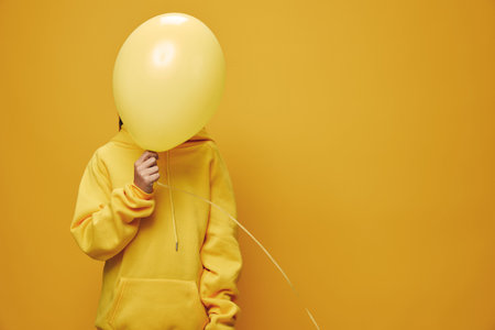 Person in yellow hoodie holding a yellow balloon covering the face standing against yellow background. Casual outfit and monochrome style with soft lighting in studio.の写真素材