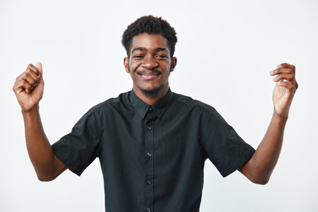 Young black man with dark skin wearing black shirt smiling with closed eyes and raised hands isolated on white background. Positive emotion and casual style portrait.の写真素材
