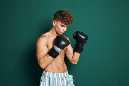 Young man with curly hair wearing boxing gloves and striped shorts training in front of green...