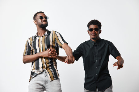 Two young black men with dark skin wearing sunglasses and casual shirts dancing and having fun together on white background. Friendship, joy and style in studio.の写真素材
