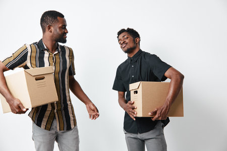 Two African American men with dark skin carrying cardboard boxes, smiling and interacting in casual clothes against white background for moving or delivery.の写真素材