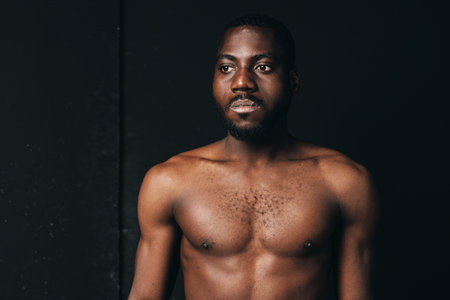 African man shirtless with muscular body and serious expression in creative conceptual visual style. Dark skin, portrait with studio lighting and black background for artistic photography.の写真素材