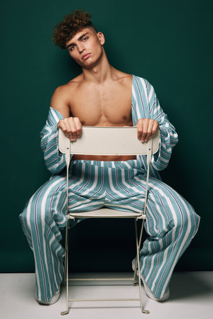 Young man with curly hair wearing striped pajamas sitting backwards on white chair against dark green background in studio portrait fashion concept.の写真素材