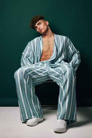 Young man with curly hair wearing striped loose suit and white sneakers sitting on chair against dark background. Fashion model posing with open shirt and casual style in studio.の写真素材