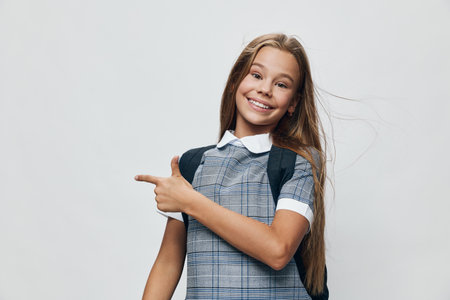 Smiling schoolgirl with long hair wearing checkered dress and backpack pointing with finger on light background. Happy child, education, casual and cheerful girl with positive pose.の写真素材