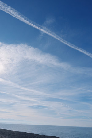 Blue sky over calm sea and pebbled beach with wispy clouds and contrails, a tranquil coastal seascape at the horizon.の写真素材