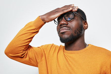 African man with dark skin and beard wearing glasses and orange sweater holding forehead with closed...