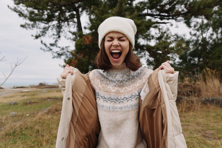 Woman in a cozy sweater and beanie on a windy outdoor day, laughing and pulling open a light coat, framed by an open field and trees in a natural landscape.の写真素材