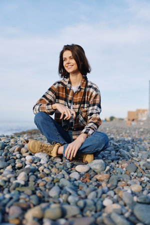 Smiling woman sits on a pebble beach with a camera hanging at her neck, wearing a plaid casual shirt...